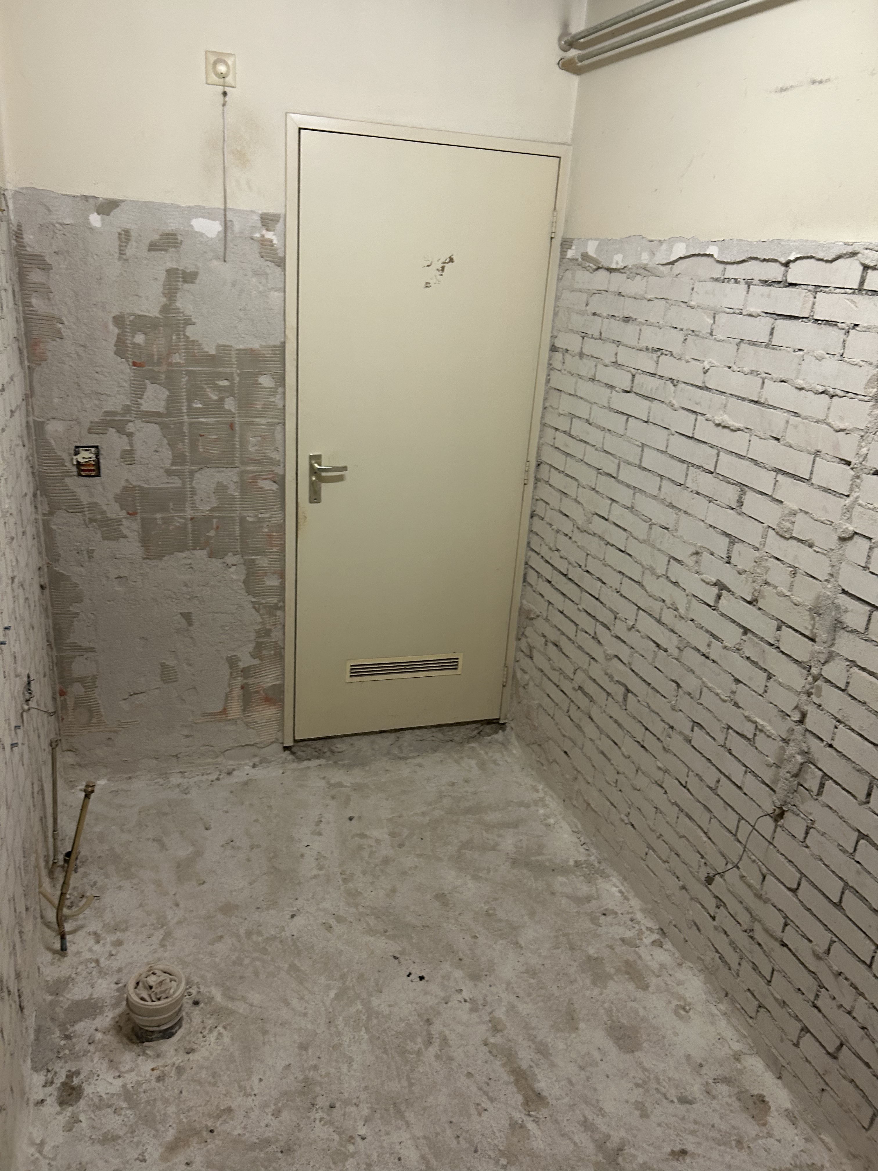 Tweede aanzicht van dezelfde gestript badkamer, leidingen en vloer zichtbaar.